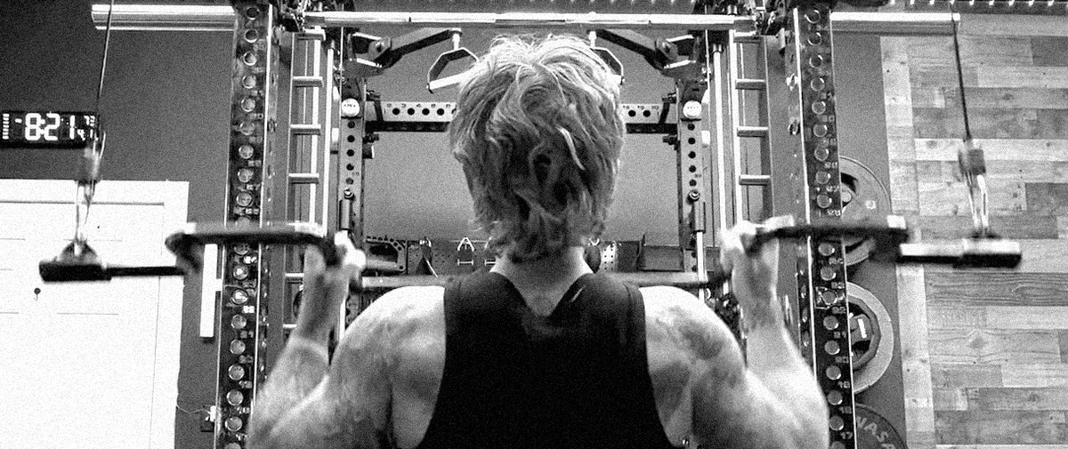 A black and white photo of a person from behind, doing a pulldown with the Andreoni bar on a cable machine in a home gym.